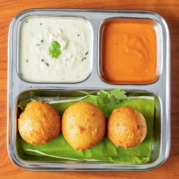 Three golden-brown medu vada on a banana leaf with white and orange chutneys in a steel tray.