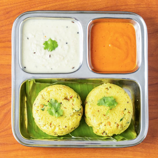 Two yellow idlis on banana leaves with white and orange chutneys in a steel thali.