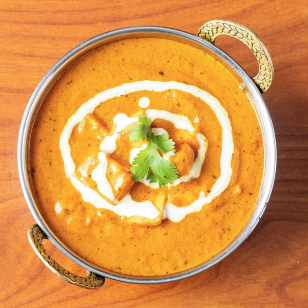 A metal bowl of orange paneer butter masala with cream swirls and a cilantro garnish.