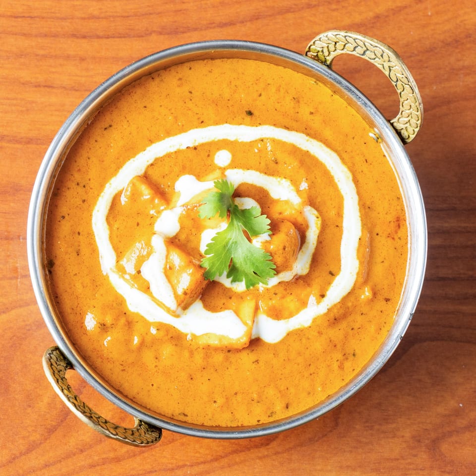 A metal bowl of orange paneer butter masala with cream swirls and a cilantro garnish.