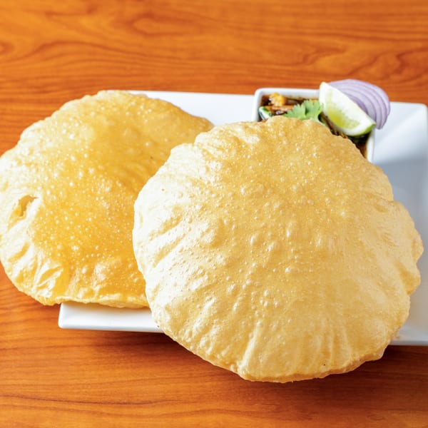 Two golden-brown puri breads on a white plate with a side of chana masala, lime, and onion.