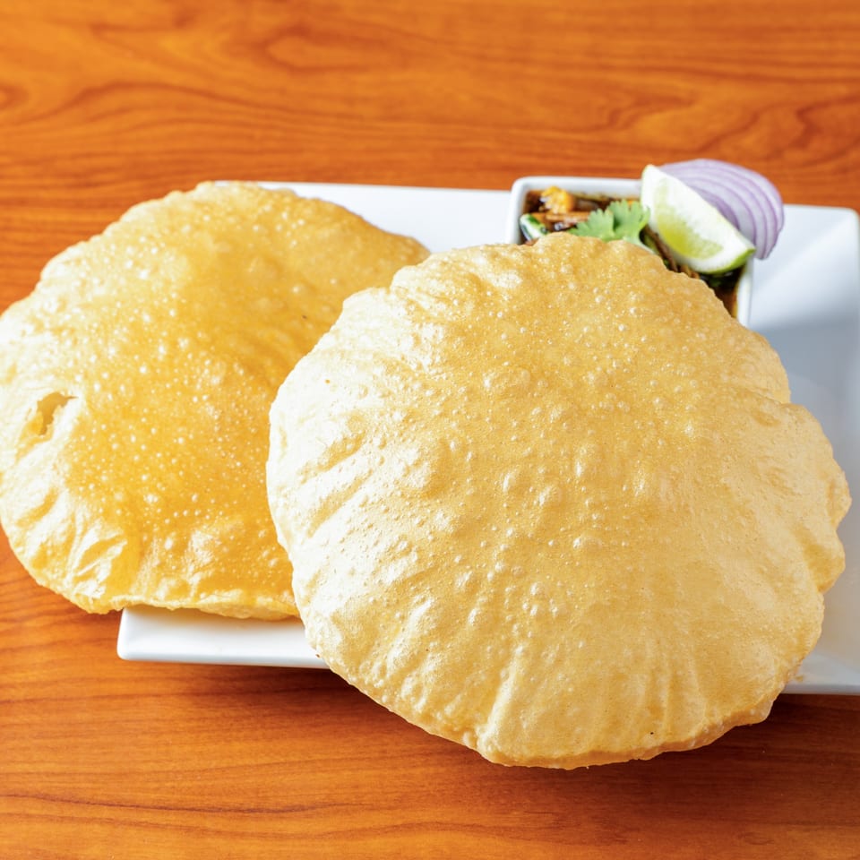 Two golden-brown puri breads on a white plate with a side of chana masala, lime, and onion.