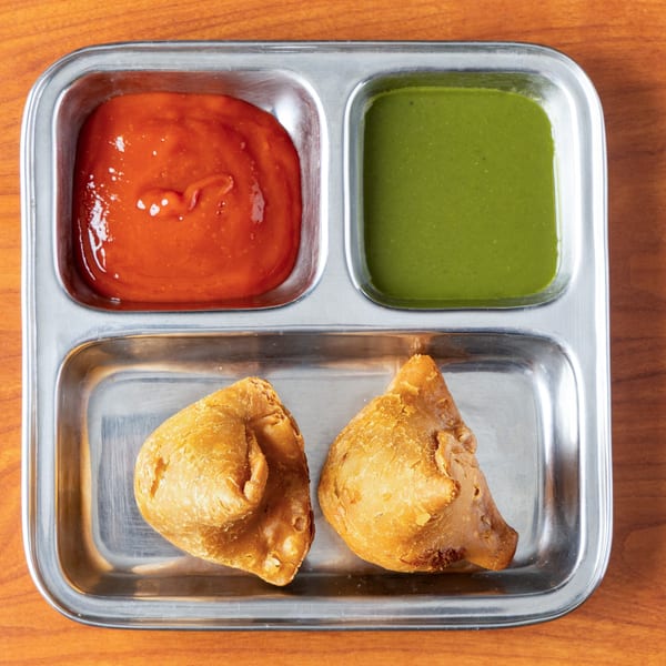 Two golden-brown samosas and two dipping sauces, red and green, on a metal tray.