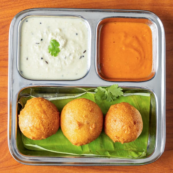 Three golden-fried medu vada on a banana leaf with coconut and tomato chutneys in a steel thali.