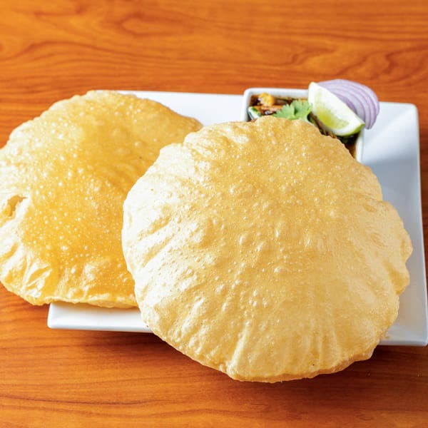 Two golden-brown puri breads on a white plate with a side of chana masala, onion, and lime.