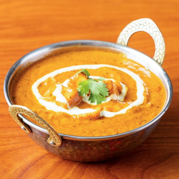 A metal bowl of orange curry with white cream swirls and a sprig of cilantro on a wooden table.