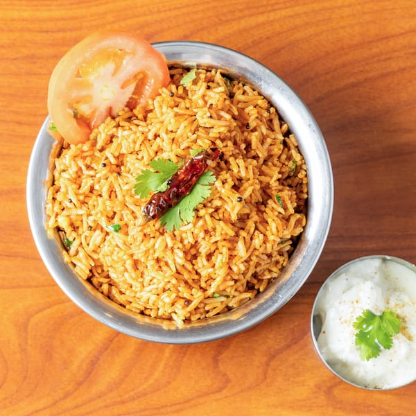 A metal bowl of tomato rice with a tomato slice, chili, and cilantro, next to a small bowl of raita.