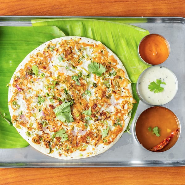 Vegetable uttapam on a banana leaf with three small bowls of sauces on a metal tray.